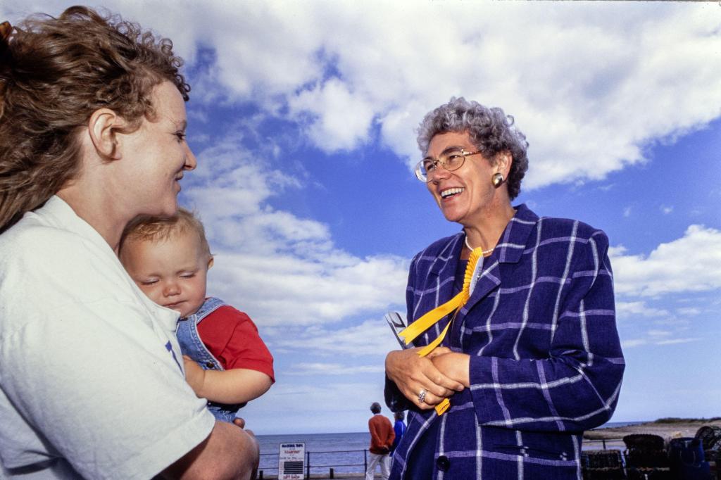Diana Maddock smiling and talking to a woman holding a baby