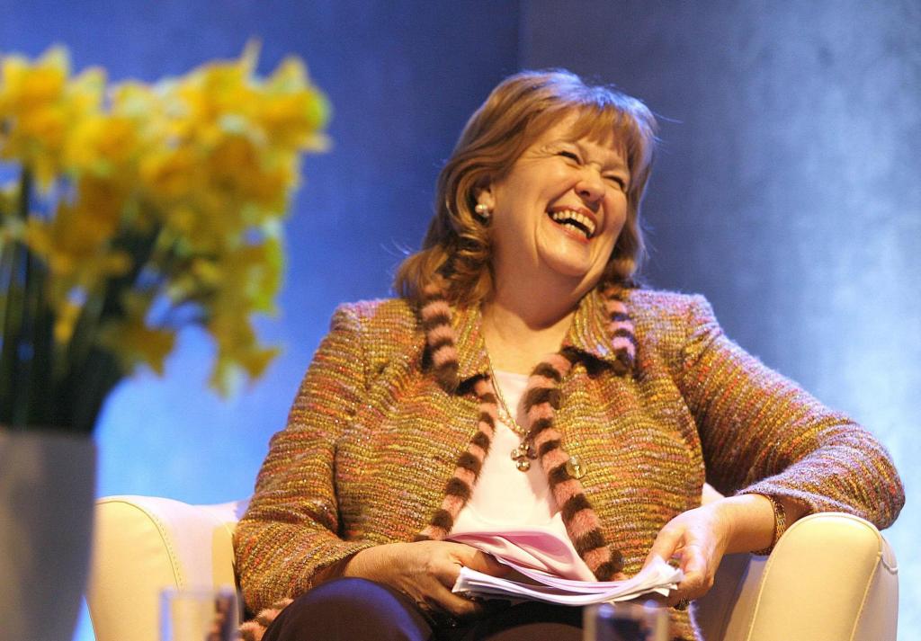 A woman sitting in a chair holding papers and laughing with a vase of daffodils in the foreground