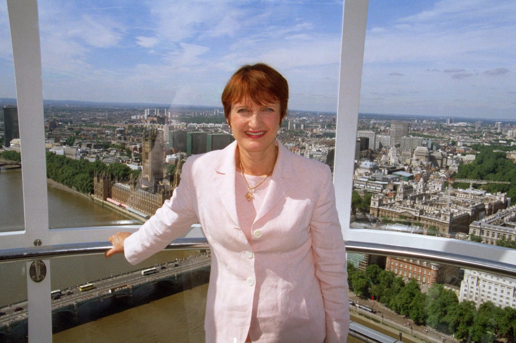 Tessa Jowell wearing a light pink suit in a London Eye capsule, with Parliament and River Thames behind