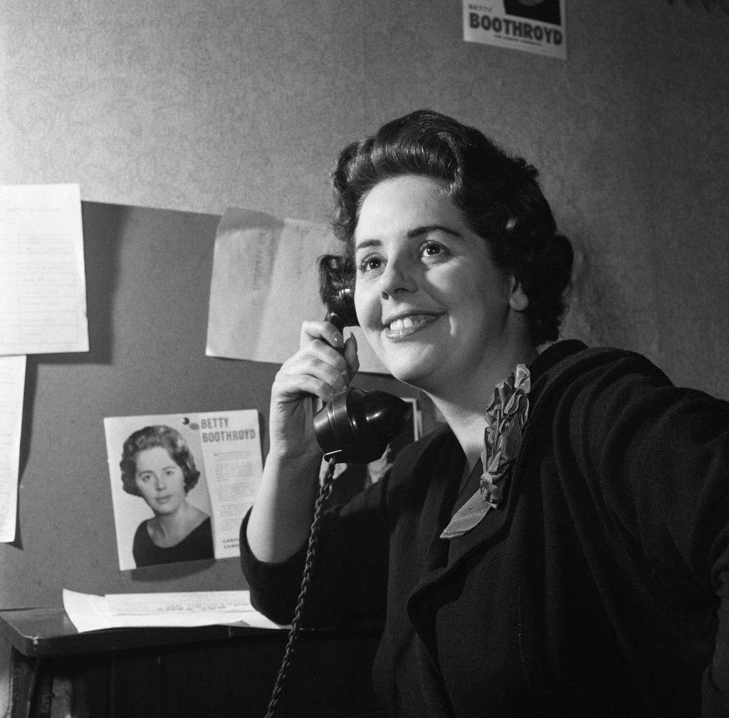 Black and white photograph of Boothroyd holding a telephone, a leaflet with her photo and name pinned on a noticeboard in the background